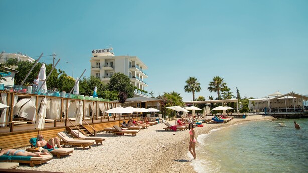 Une plage avec des chaises longues et des parasols, un bâtiment portant l’inscription « DEMI-HÔTEL » à l’arrière-plan, et un ciel bleu dégagé.