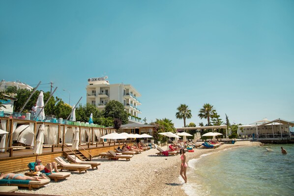 A beach with sun loungers and umbrellas, a building labeled "DEMI HOTEL" in the background, and a clear blue sky. - Demi Hotel (Sarandë)