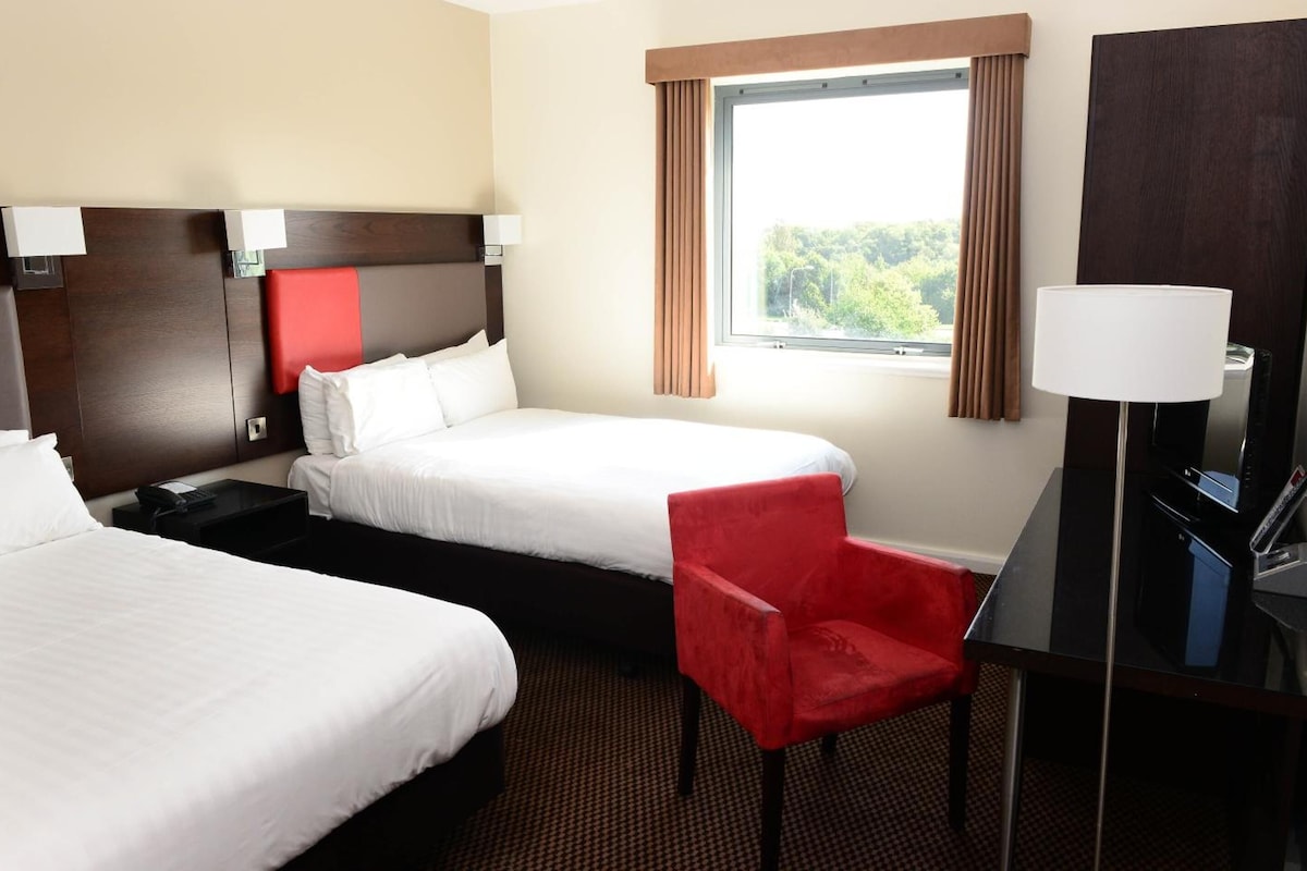 Habitación de hotel con dos camas, una silla roja, un escritorio con lámpara y una ventana con cortinas.