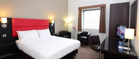 Ein Hotelzimmer mit einem großen Bett, einem Schreibtisch, einem Sessel, einem Fernseher und einer Lampe.