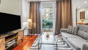 Un salon moderne doté d’un téléviseur à écran plat, d’une table basse en verre, d’un canapé gris et offrant une vue sur un balcon extérieur.