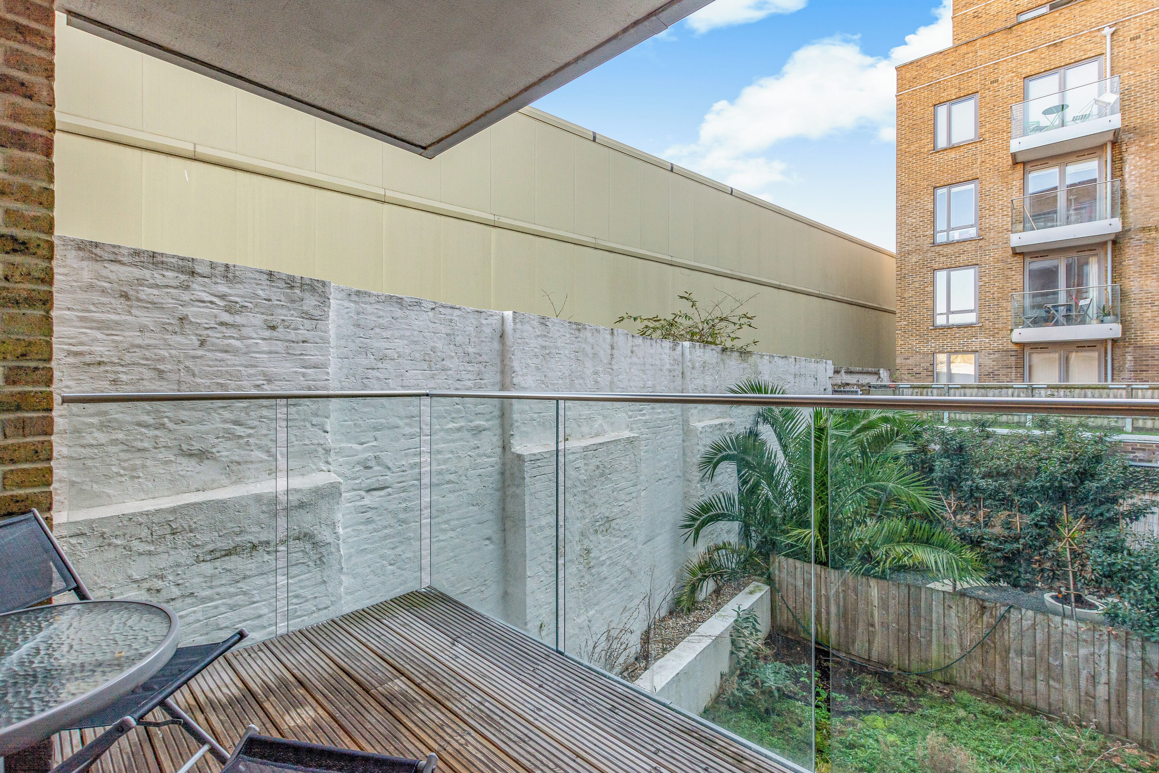 Ein Balkon mit Holzboden, Glasgeländer und Blick auf ein Backsteingebäude sowie grüne Bepflanzung.