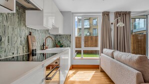 Une cuisine moderne avec des meubles blancs, un pare-feu en marbre et un plancher en bois. Une fenêtre offre une vue sur un jardin, et il y a un balcon.