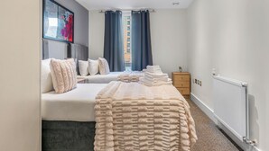Une chambre à coucher moderne avec un lit, une grande fenêtre avec des rideaux, un radiateur et une petite armoire en bois.