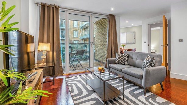 Un salon moderne avec un canapé gris, une table basse en verre et un tapis à motifs zébrés. Une porte-fenêtre coulissante donne accès à un espace extérieur doté d’un mur en briques et d’un balcon.