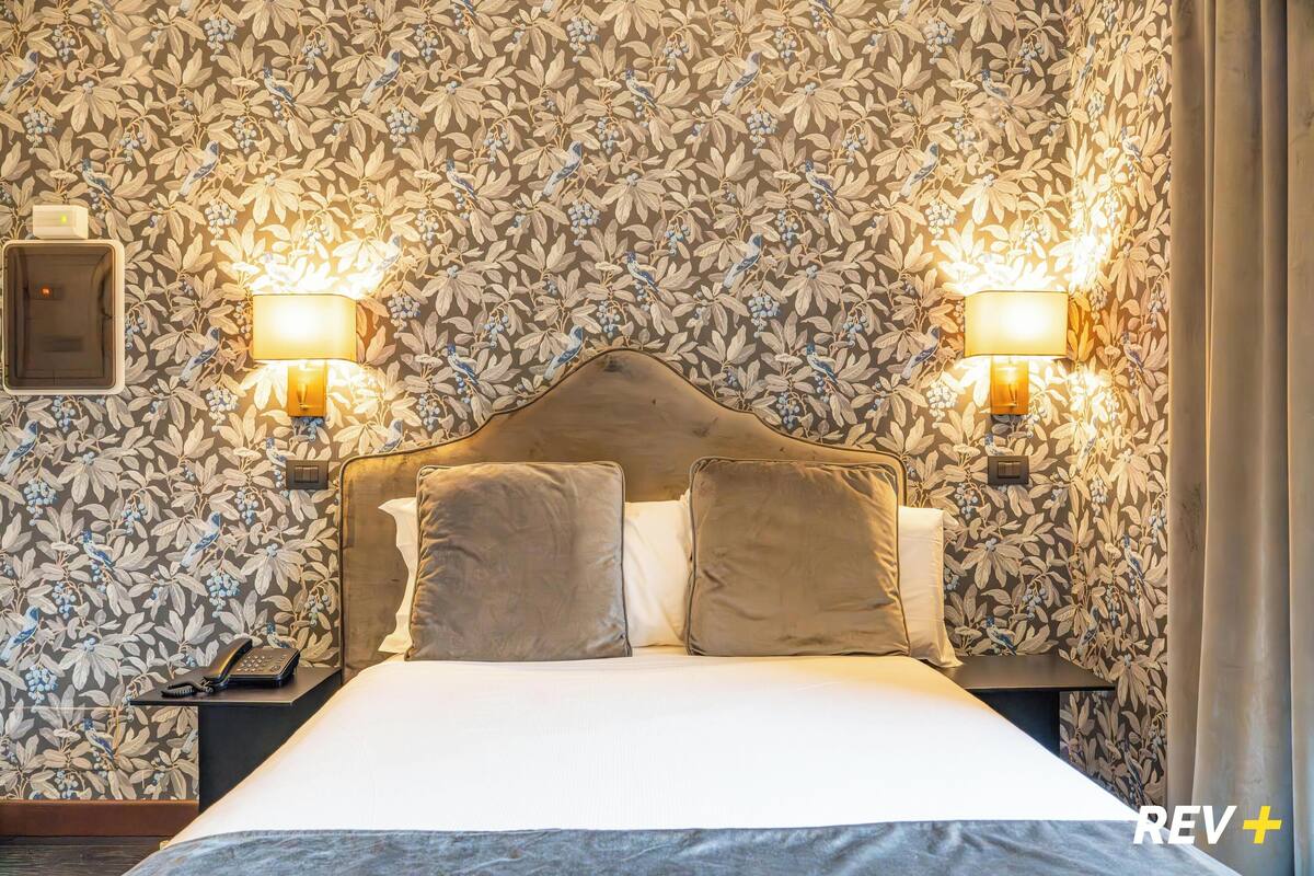 A hotel room with a floral wallpaper, two bedside lamps, a telephone, and a bed with two pillows.