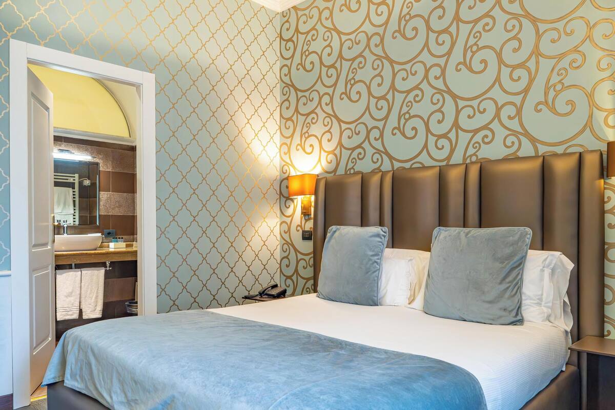 A hotel room with a large bed, a bedside table, a nightstand, a wall-mounted lamp, and a bathroom visible through an open door.