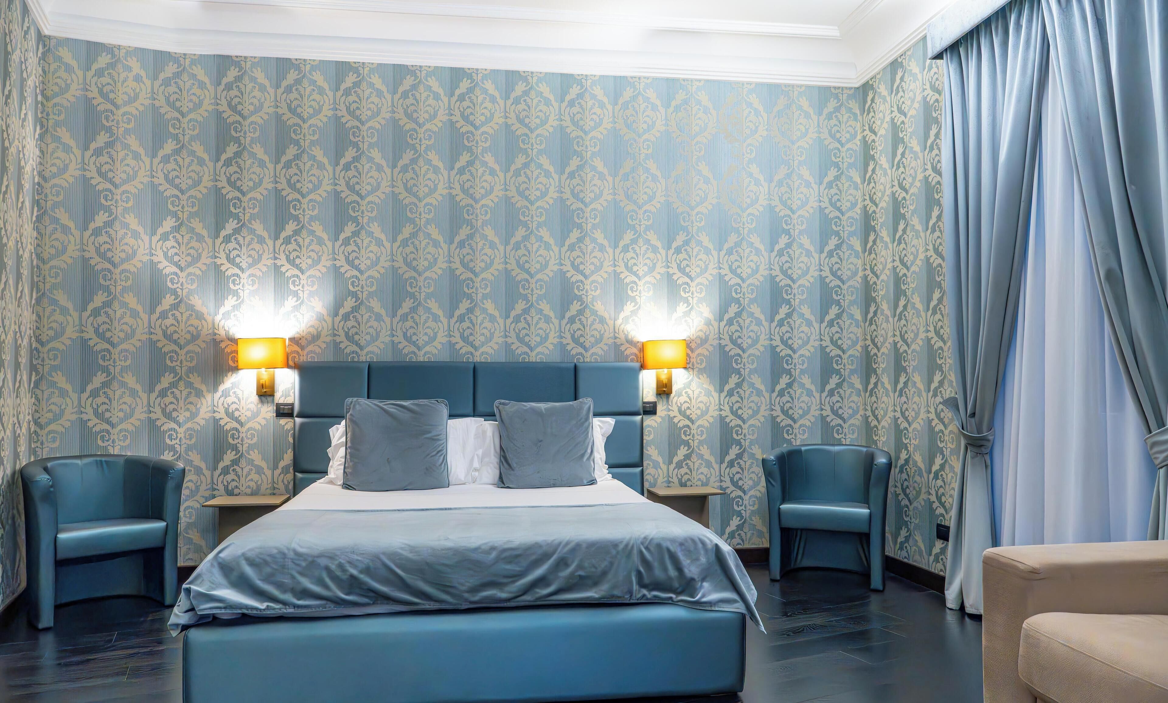 A hotel room with a large bed, two bedside tables with lamps, a blue headboard, and patterned wallpaper.