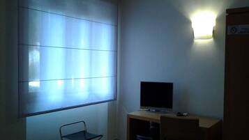 A small room with a desk, a chair, a television, and a window with blinds.