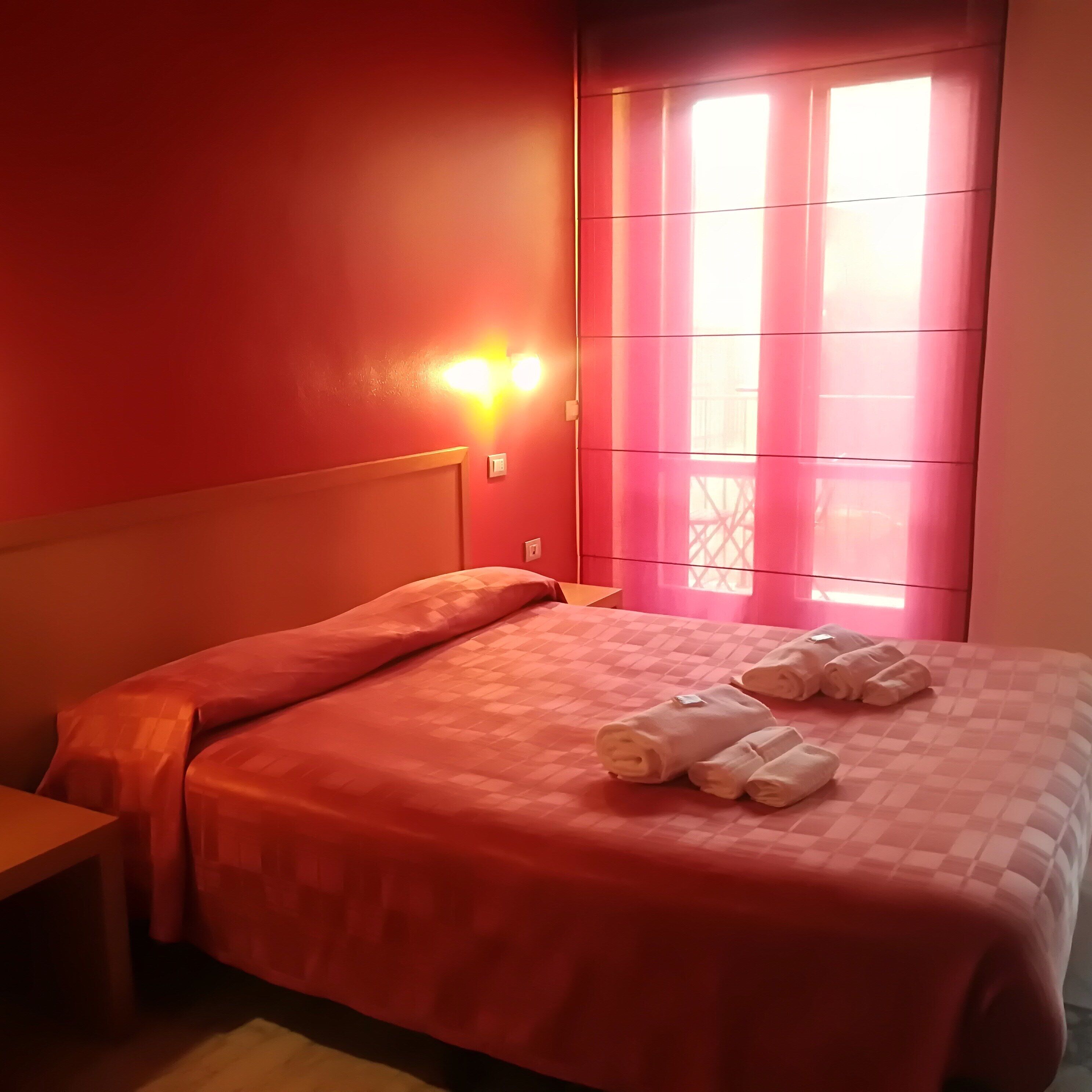 A hotel room with a red wall, a bed with a checkered bedspread, and a window with pink curtains.