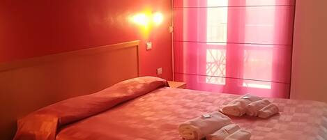 A hotel room with a red wall, a bed with a checkered bedspread, and a window with pink curtains.