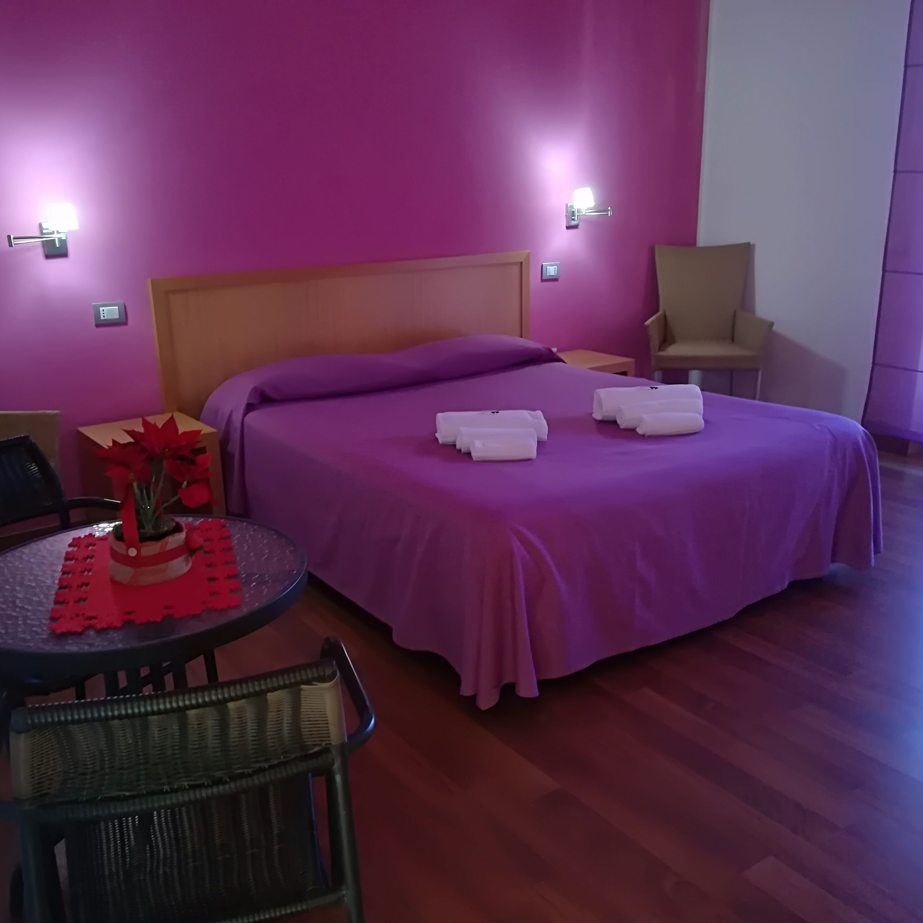 Habitación de hotel con cama, una silla, una mesita con una planta, y una lámpara fijada a la pared.