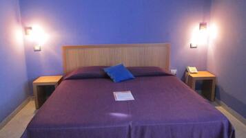 Una cama bien hecha con una colcha morada y una almohada azul, dos mesitas de noche con lámparas, y un cabecero de madera.
