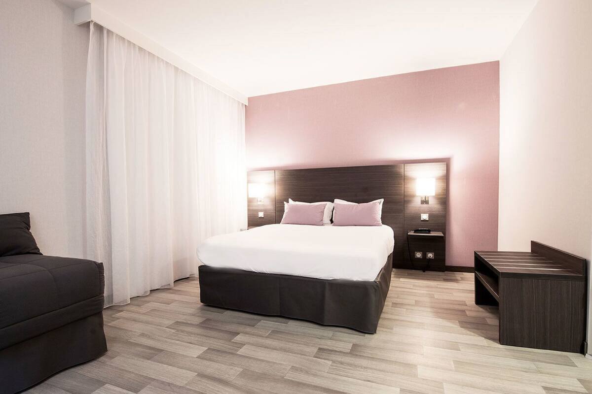 Quarto de hotel moderno com uma cama grande, um sofá escuro, uma mesinha de cabeceira e um criado-mudo.
