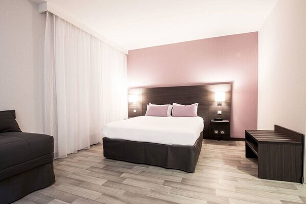 Une chambre d’hôtel moderne avec un grand lit, un canapé sombre, une table de chevet et une petite table de nuit.
