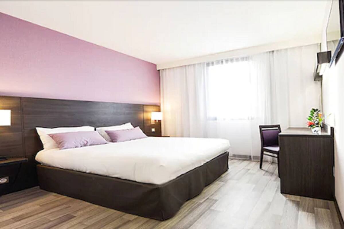 Quarto de hotel com uma cama grande, mesas de cabeceira com luminárias, uma escrivaninha com cadeira e uma janela com cortinas.