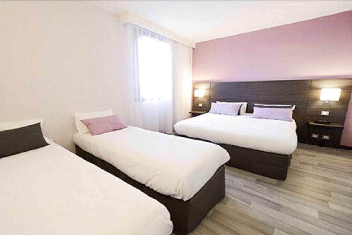 Quarto de hotel com duas camas de solteiro e uma cama de casal, roupa de cama branca, piso de madeira e uma janela com cortinas.