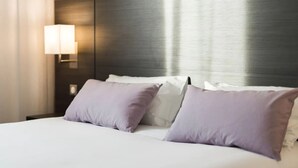 A neatly made bed with white and lavender pillows, a dark headboard, and a wall-mounted lamp. - Hôtel Eurocentre Toulouse Nord 3 Stars (Castelnau-d'Estretefonds)