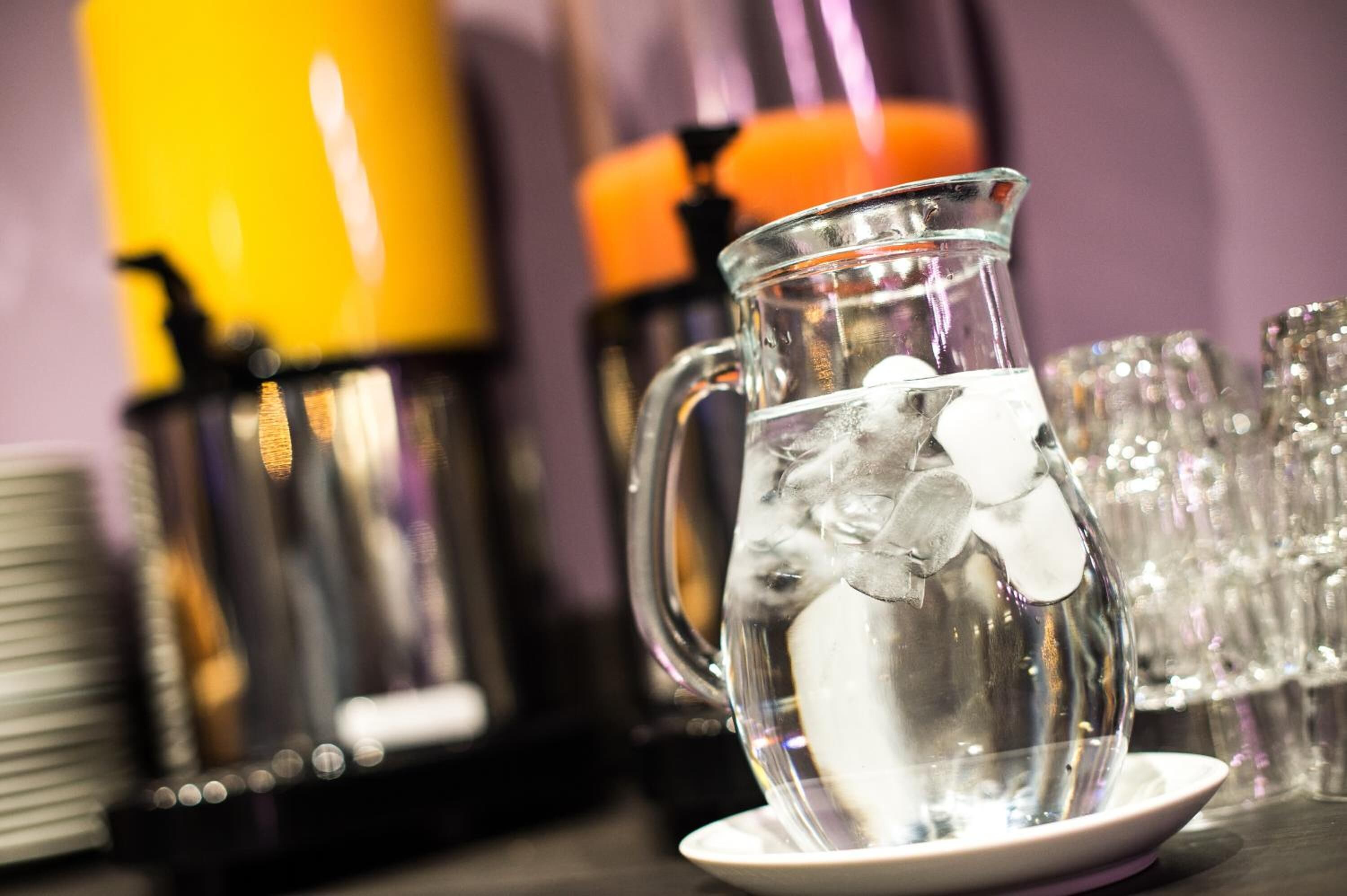 Une carafe en verre transparent contenant des glaçons, posée sur une assiette blanche, entourée d’assiettes empilées et d’un récipient jaune en arrière-plan.