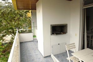 Un balcón con piso de baldosas, una silla blanca, una mesa y una parrilla empotrada.