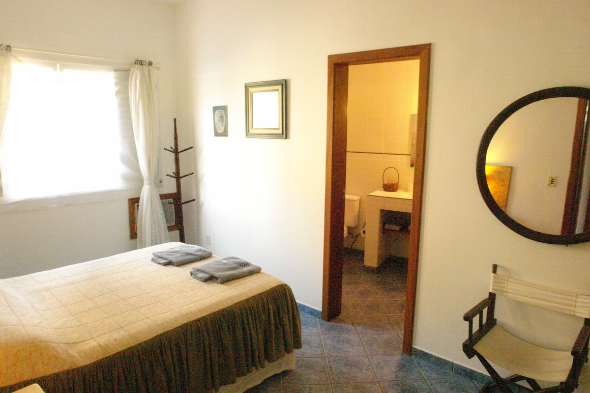 Une chambre à coucher avec un lit, une chaise, un miroir et une salle de bain visible à travers une porte ouverte.