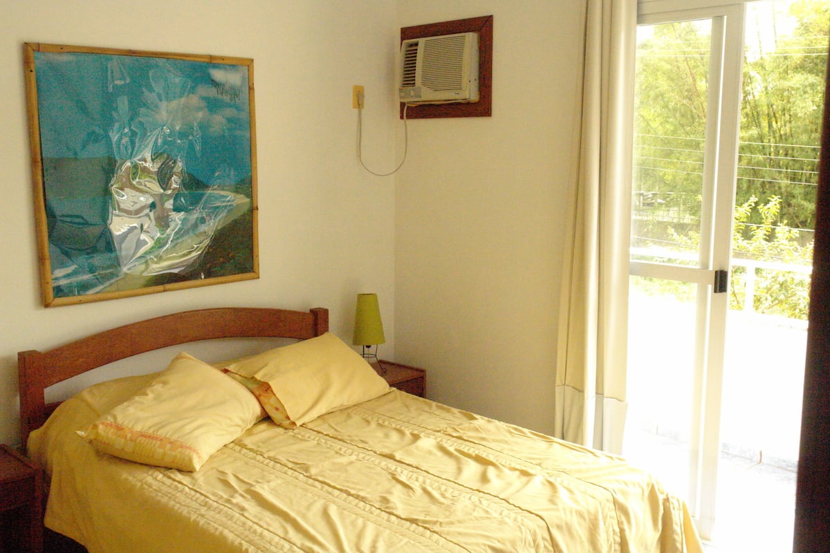Une chambre à coucher comprenant un lit, un tableau, un climatiseur, une table de chevet et une fenêtre donnant sur un paysage verdoyant.