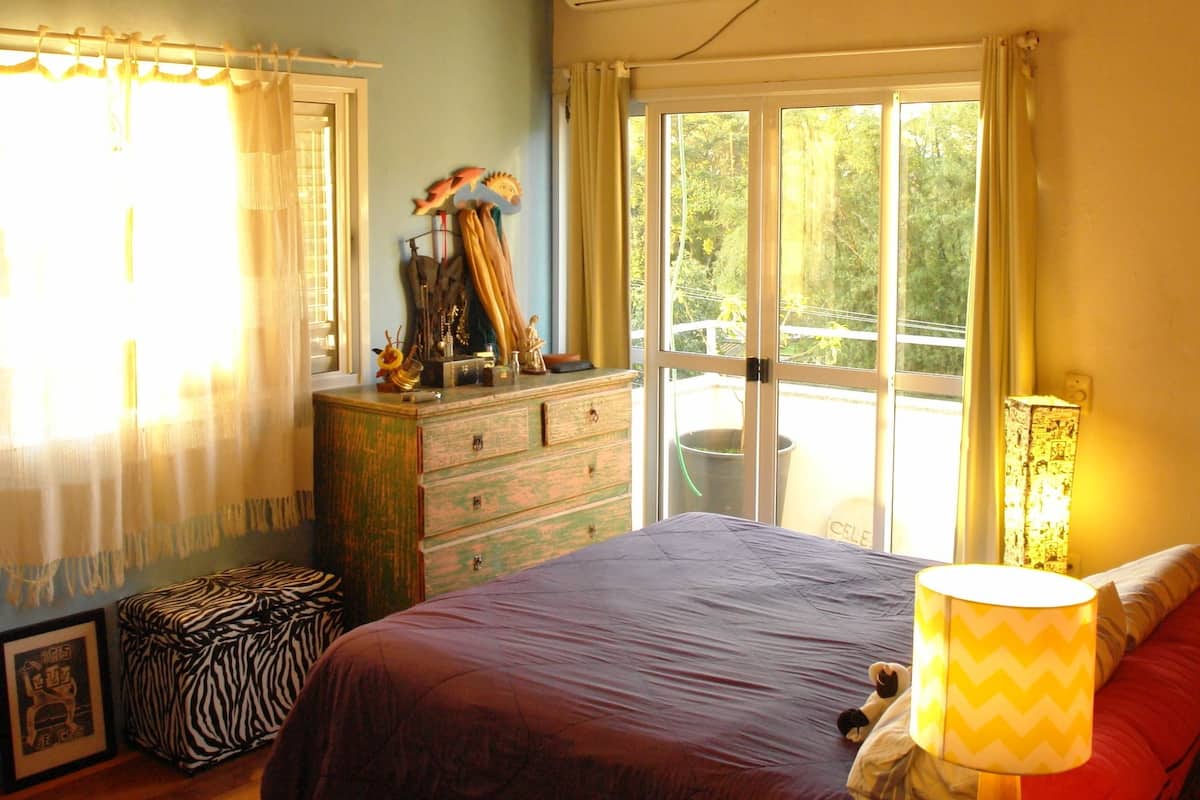 Une chambre à coucher comprenant un lit, une commode, un ventilateur de plafond, une fenêtre avec des rideaux et une table de chevet avec une lampe.