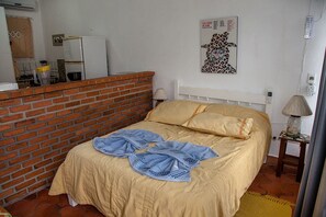 Un dormitorio con separador de ladrillo, una cama con ropa de cama color beige, un cabecero blanco, un cartel en la pared y una mesita de noche con lámpara.
