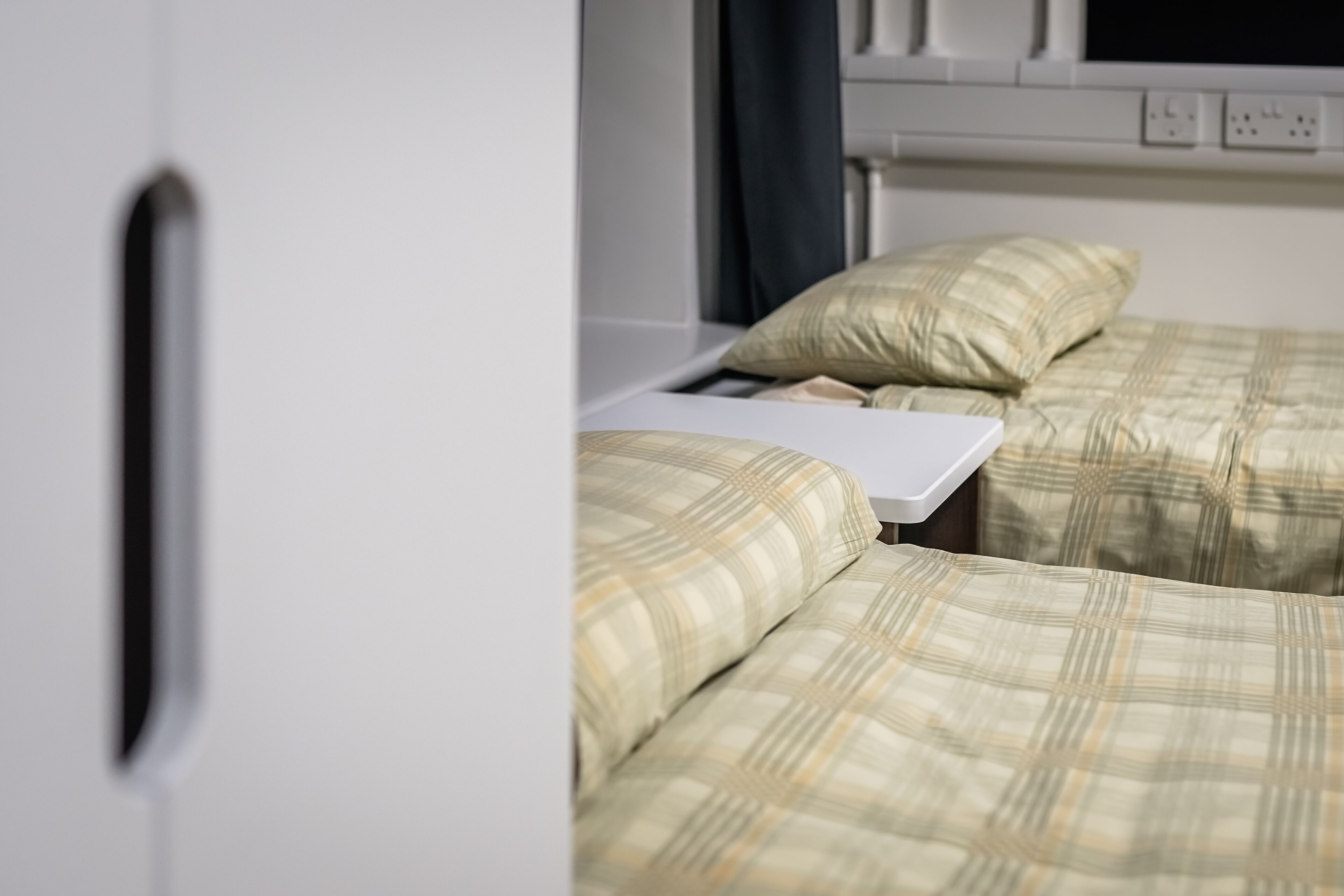 A bedroom with two beds, each with a plaid duvet cover, a white bedside table, and a window with a view.