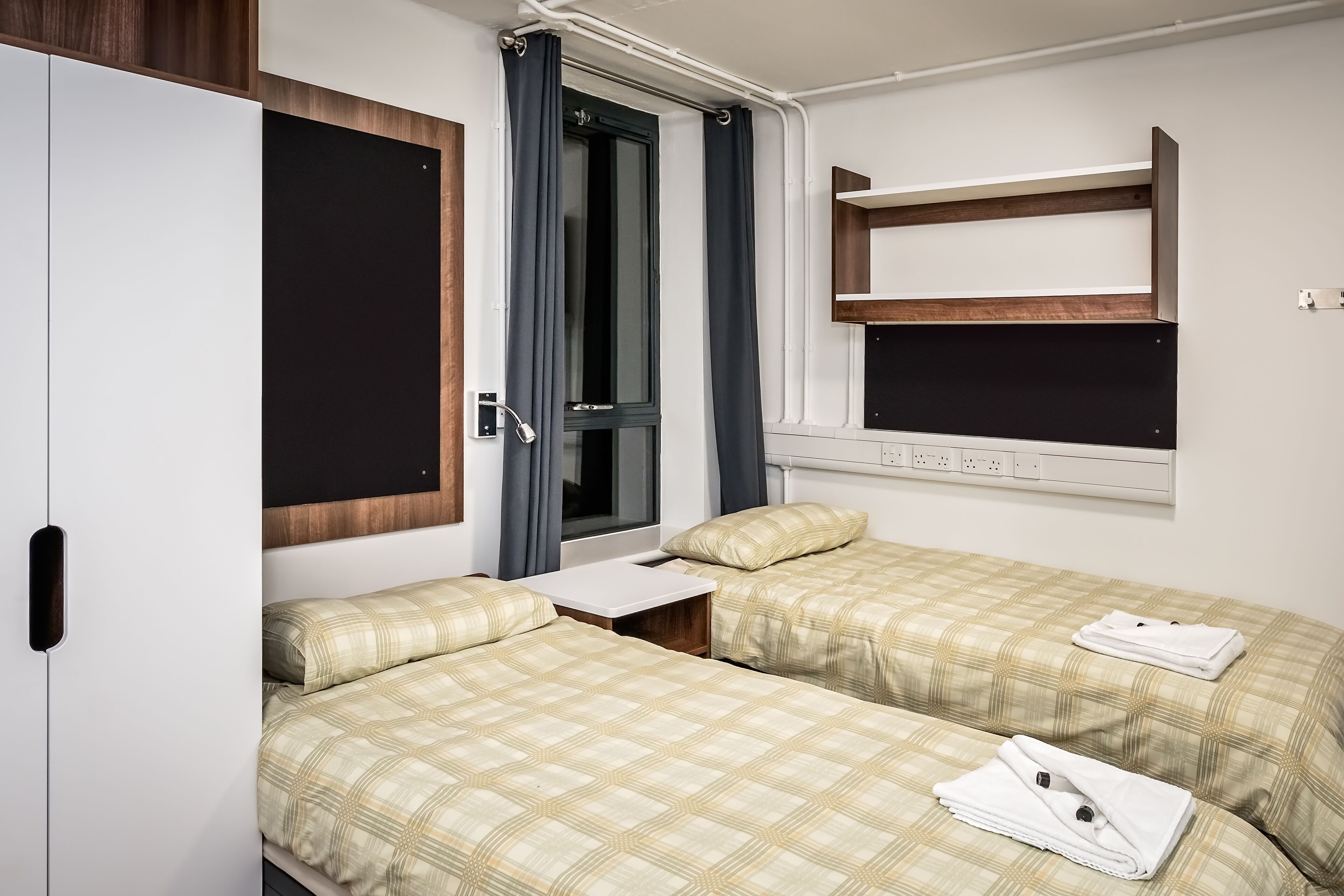 A bedroom with two beds, a wardrobe, a window with curtains, and a shelf above the bed.