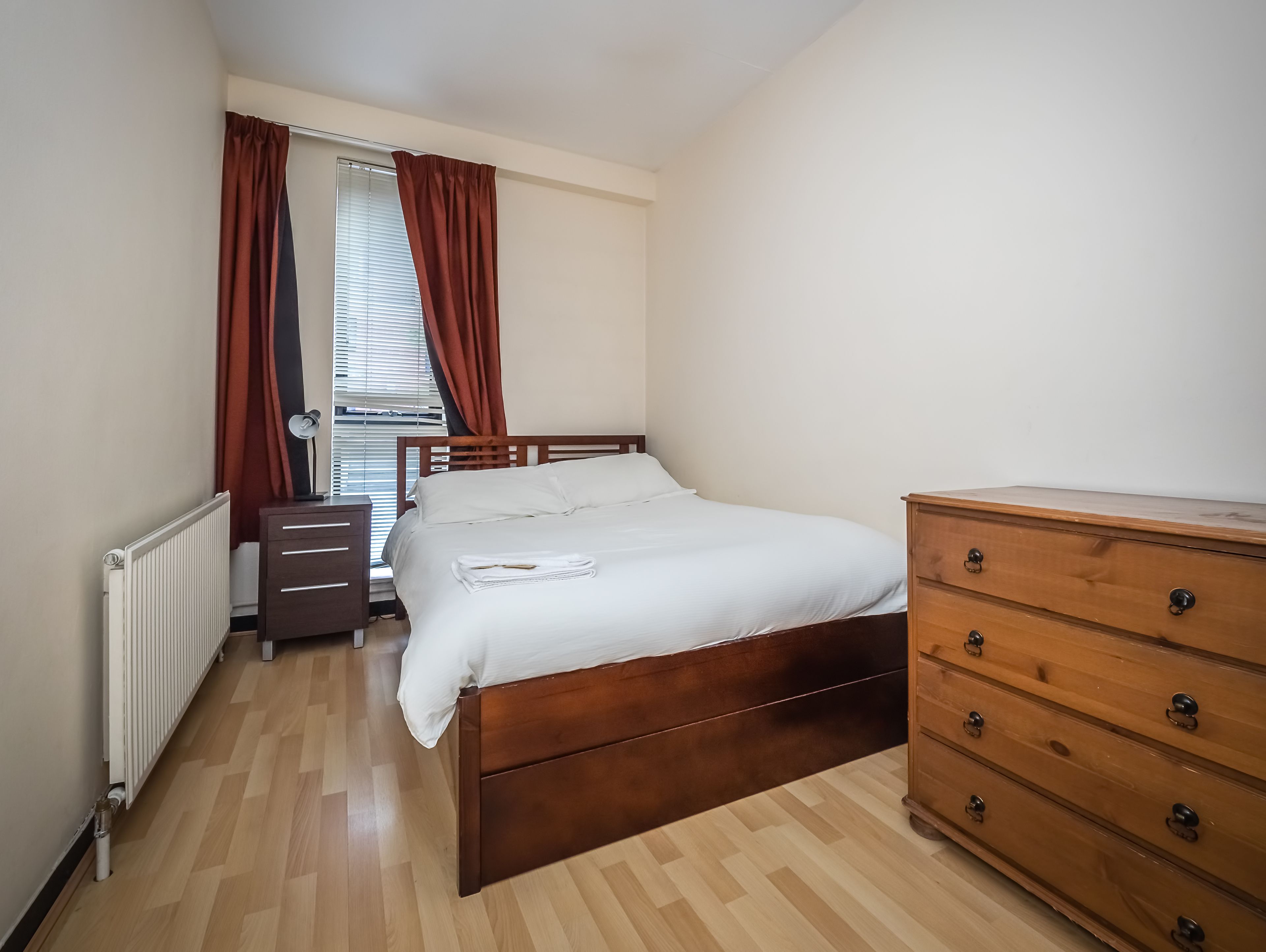 Quarto com cama de madeira, cómoda de madeira, radiador e janela com cortinas.
