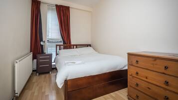 Quarto com cama de madeira, cómoda de madeira, radiador e janela com cortinas.