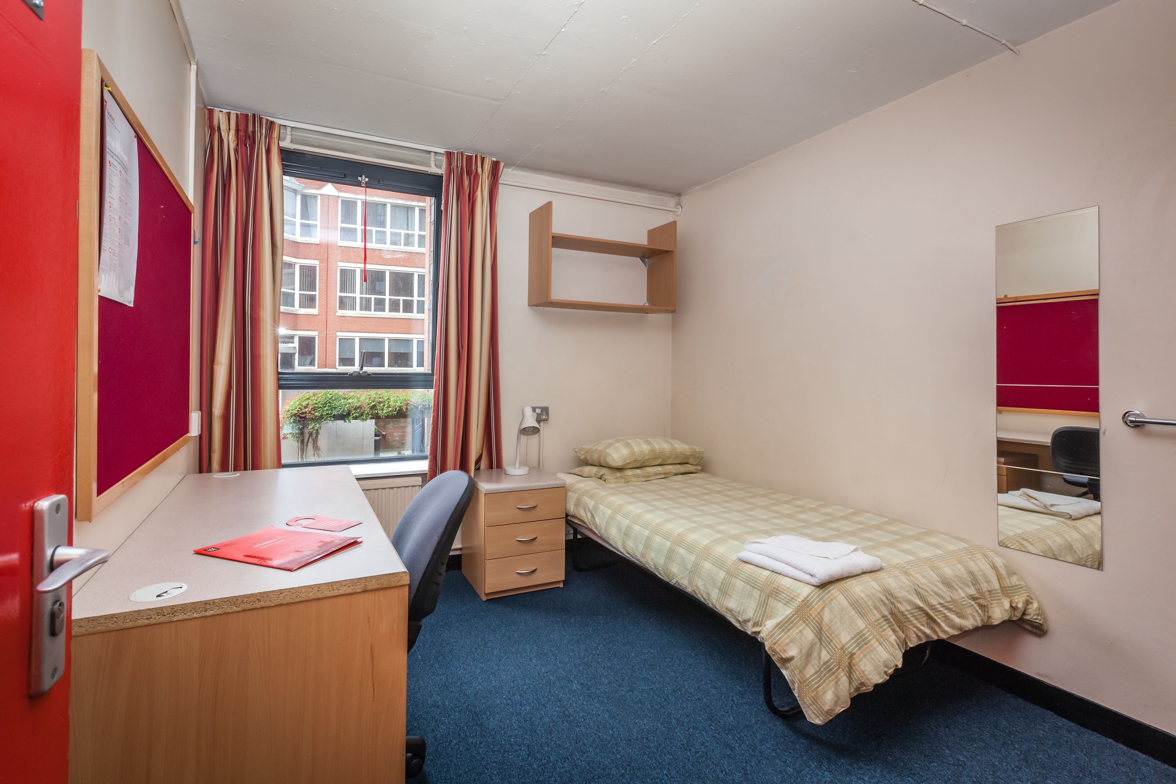 A small, tidy room with a single bed, a desk, a chair, and a window with curtains.