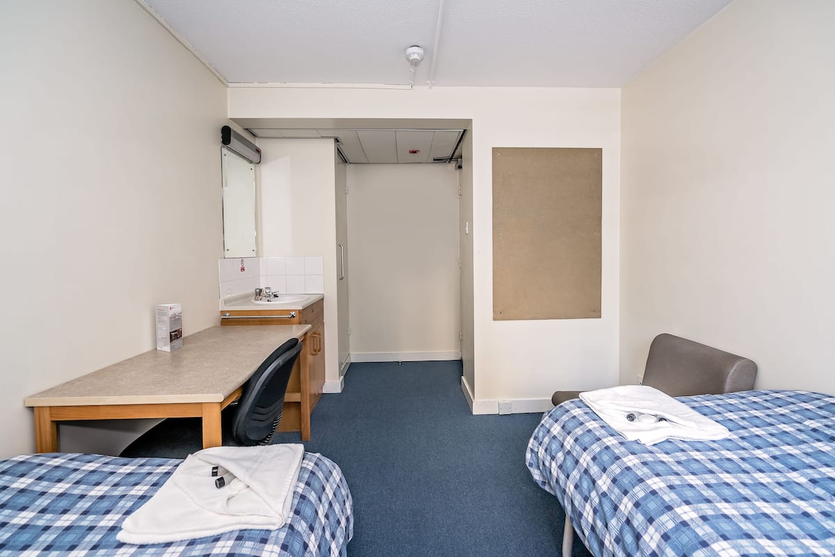 Une petite chambre propre, équipée de deux lits, d’un bureau, d’une chaise et d’une salle de bain.
