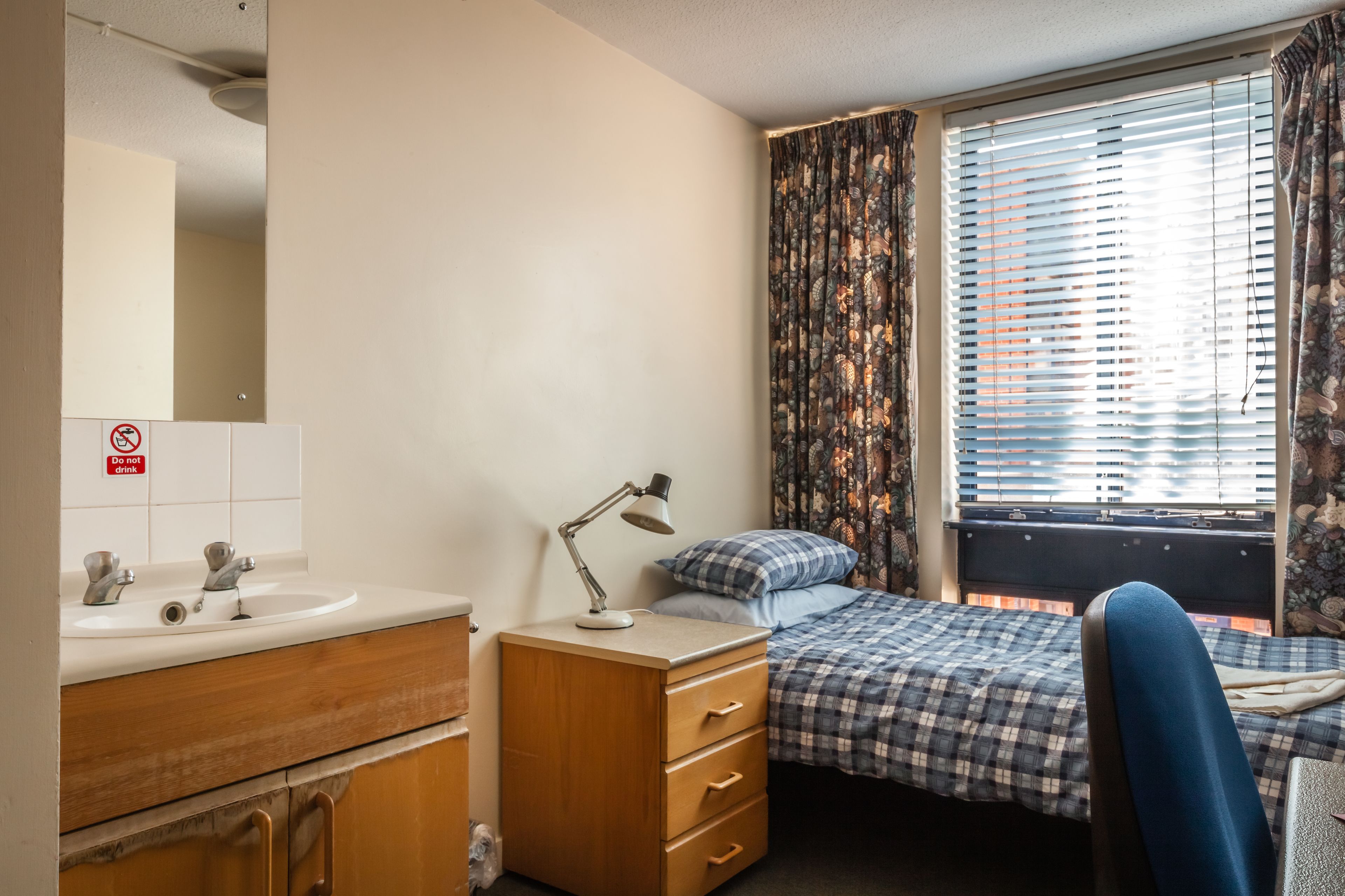 Ein Schlafzimmer mit Bett, Schreibtisch, Stuhl, einem Fenster mit Vorhängen und einem Badezimmer mit Waschbecken und Spiegel.