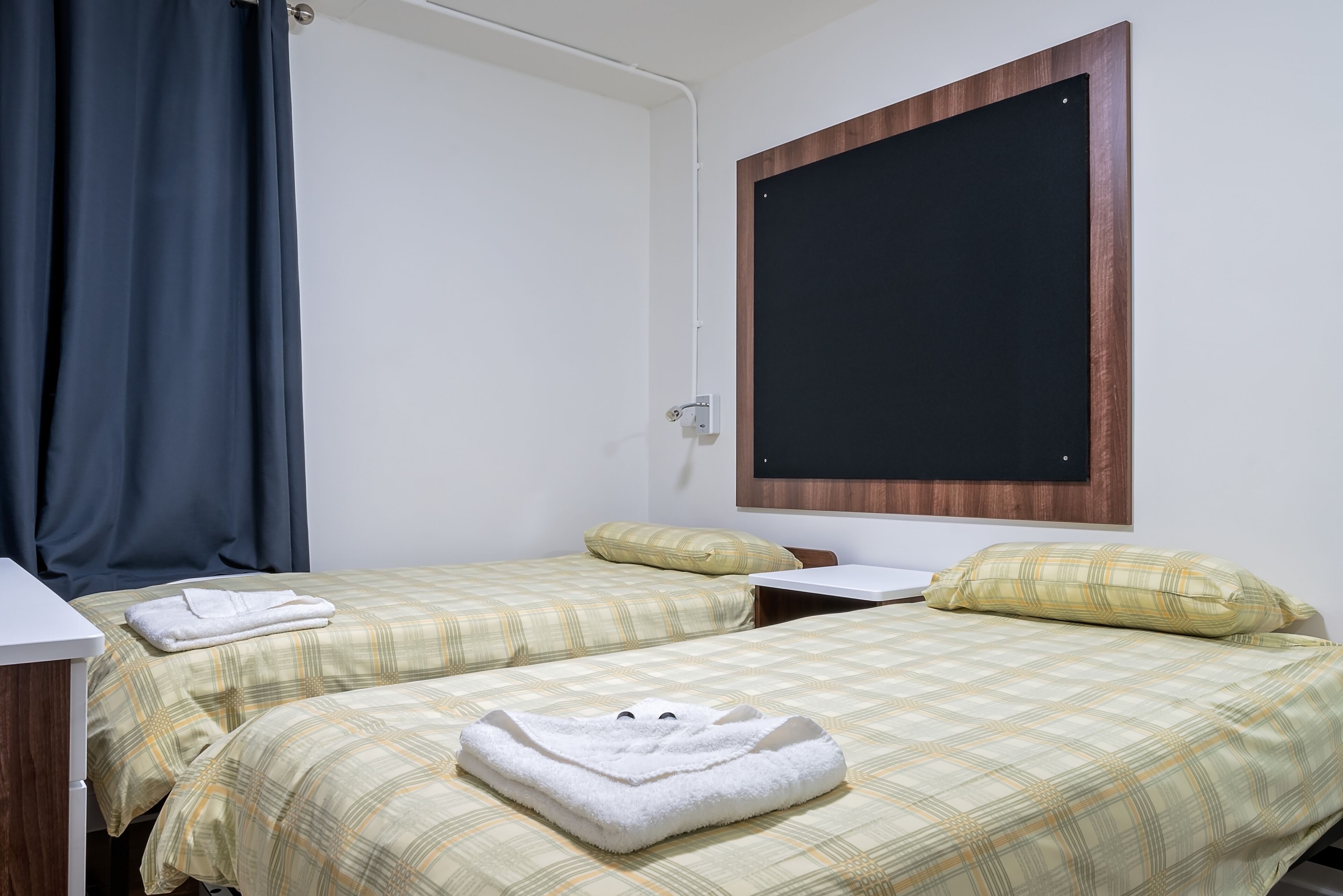 Ein Zimmer mit zwei Betten, jeweils mit einer karierten Bettdecke und Kissen, einem weißen Nachttisch und einer Tafel an der Wand.