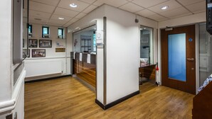 Un área de recepción con piso de madera, paredes blancas y un mostrador de recepción. Hay cuadros enmarcados en la pared, una pizarra de anuncios y una puerta azul.