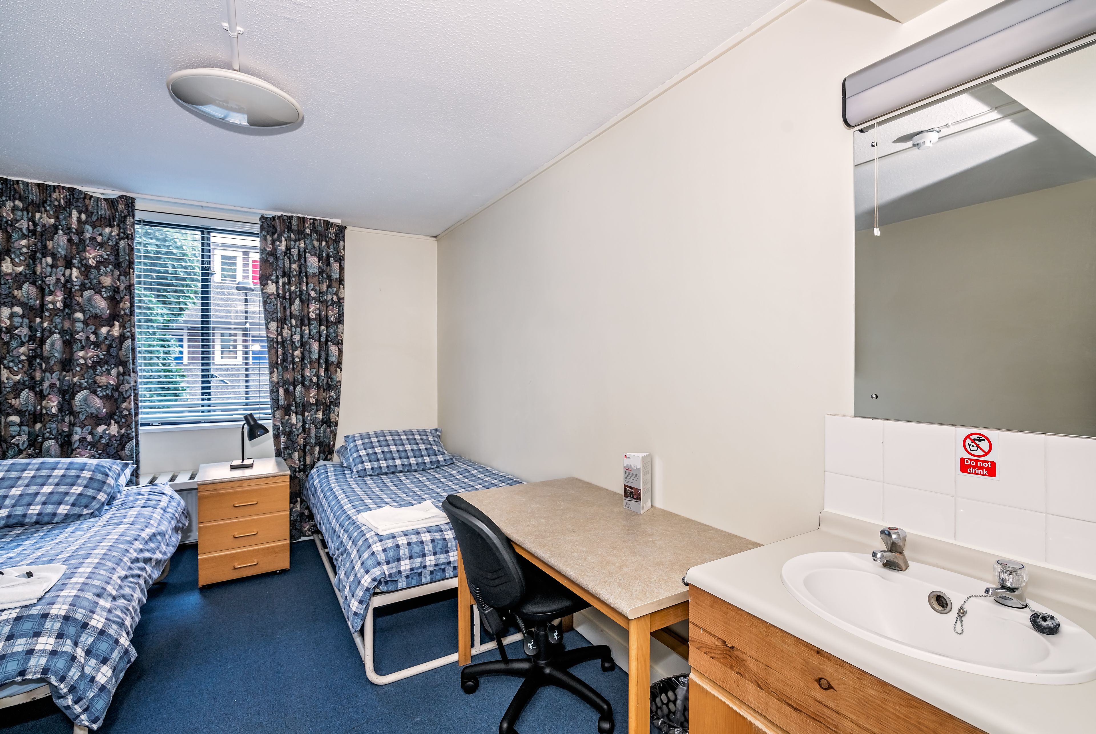 A dormitory room with two single beds, a desk, a chair, a sink, and a window with curtains.
