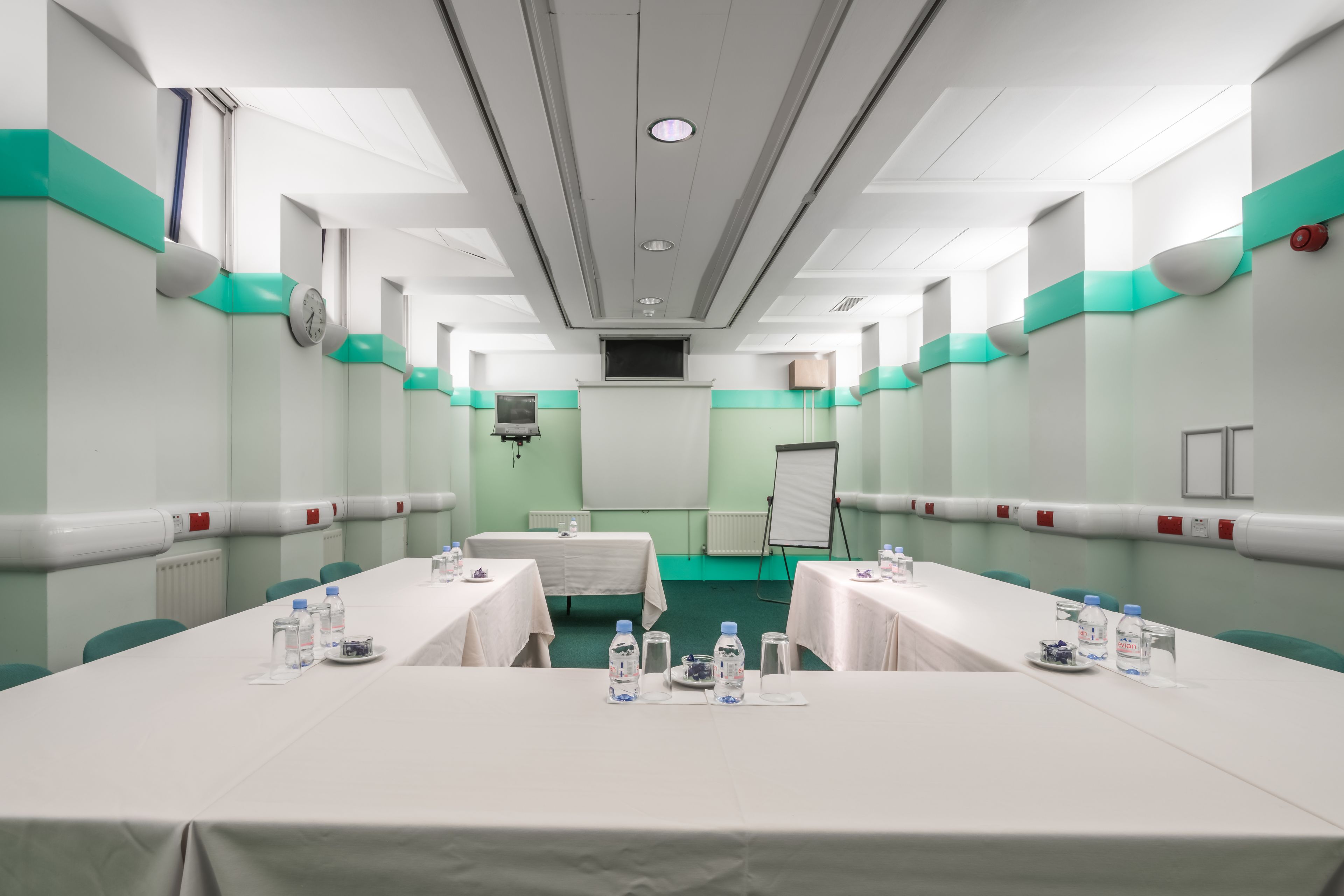 A conference room with a long table, chairs, a clock, a whiteboard, and a projector screen.