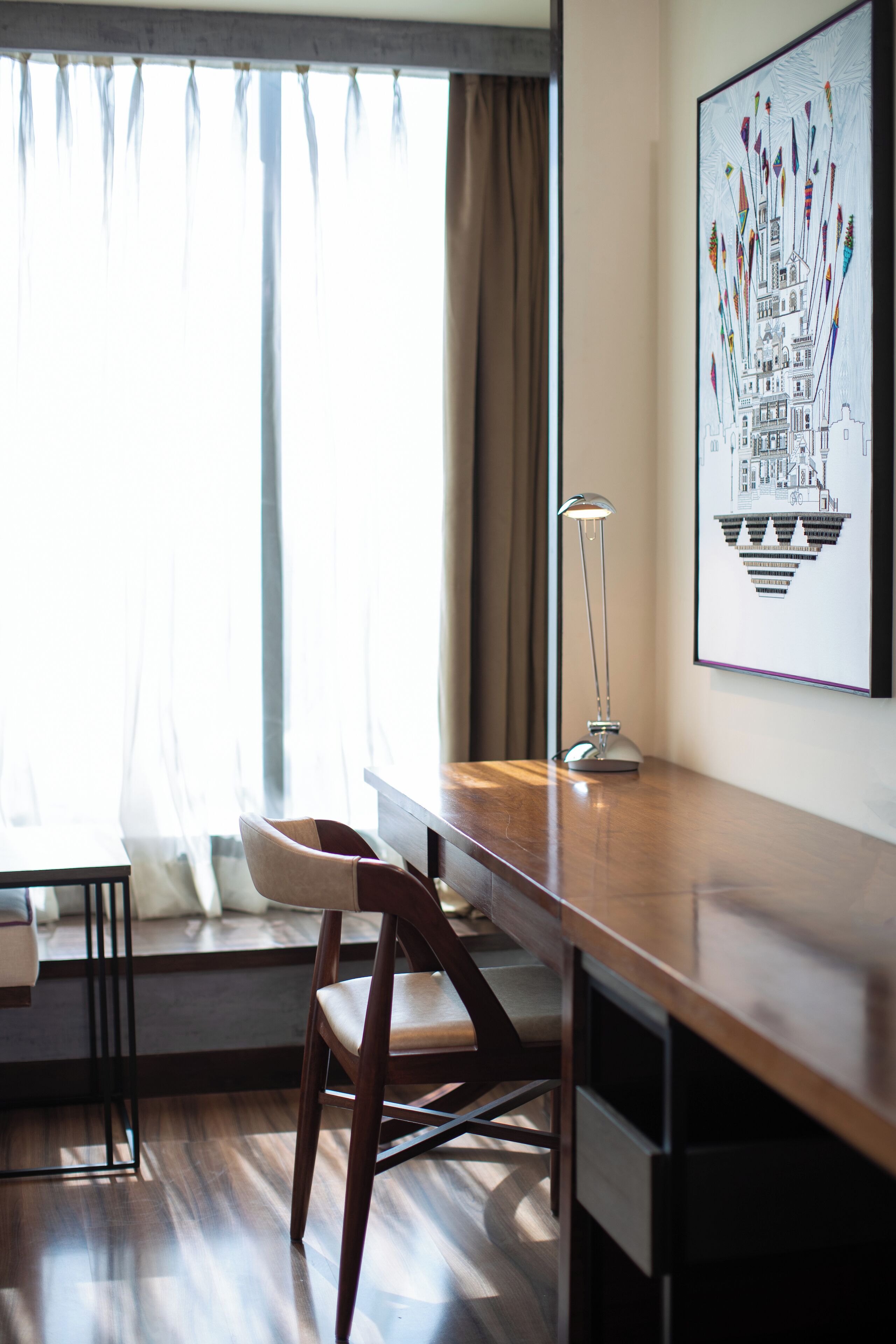 A modern room with a wooden desk, a chair, a desk lamp, and a large window with curtains.