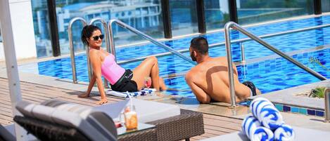 Un área junto a la piscina con sillones de descanso, una piscina con mosaicos azules y vistas a la ciudad a través de grandes ventanales.