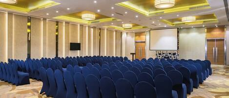 A conference room with rows of blue chairs, a large screen, and a podium.
