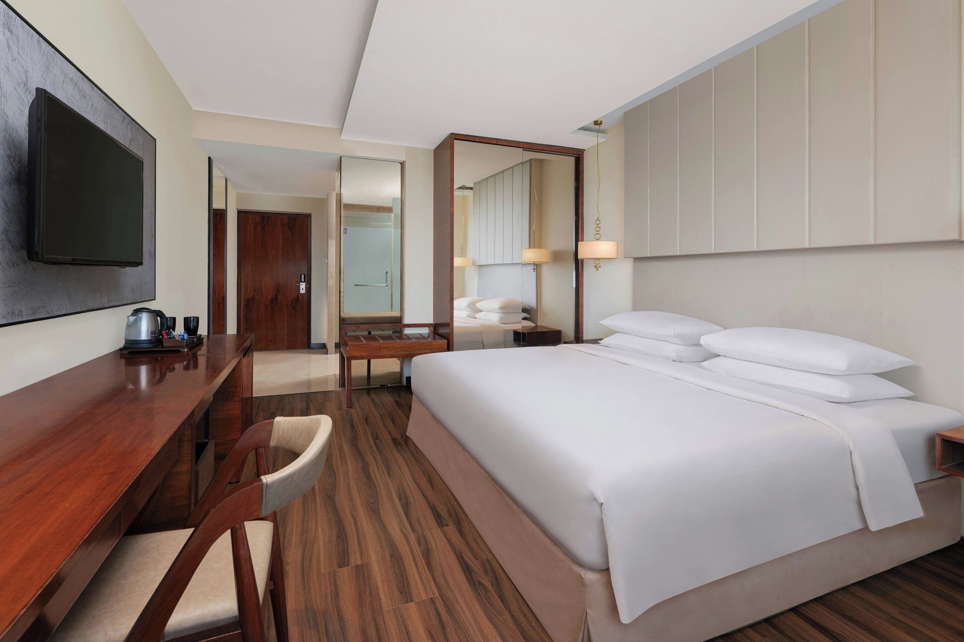 A hotel room with a large bed, a desk with a chair, a television, a mirror, and a bathroom visible through an open door.