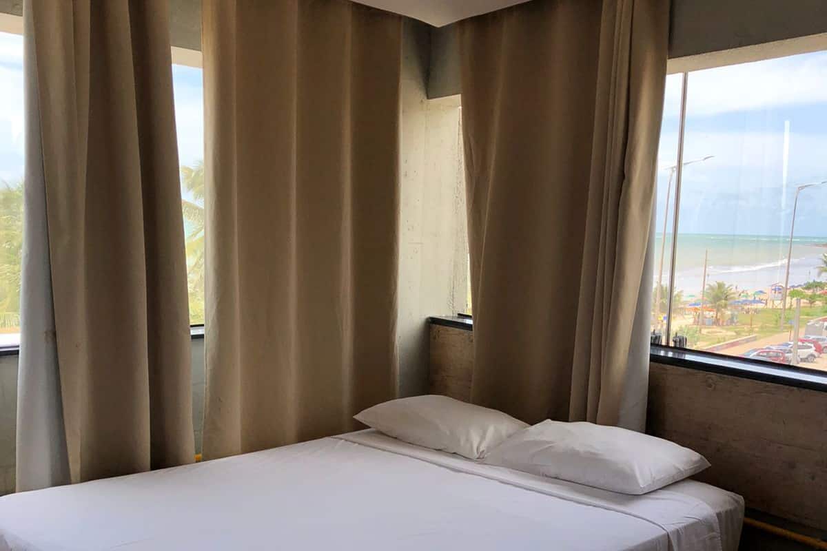 A single bed with white bedding, beige curtains, and a view of the beach through a window.