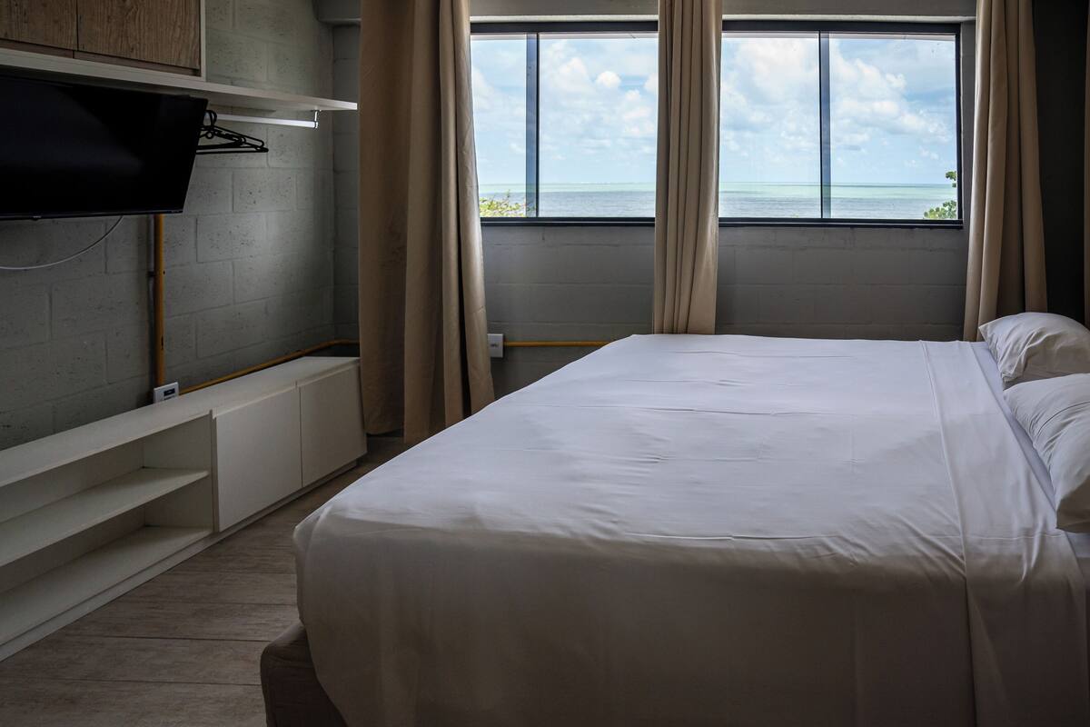 A bedroom with a bed, a TV mounted on the wall, a window with a view of the sea, and a built-in bench.