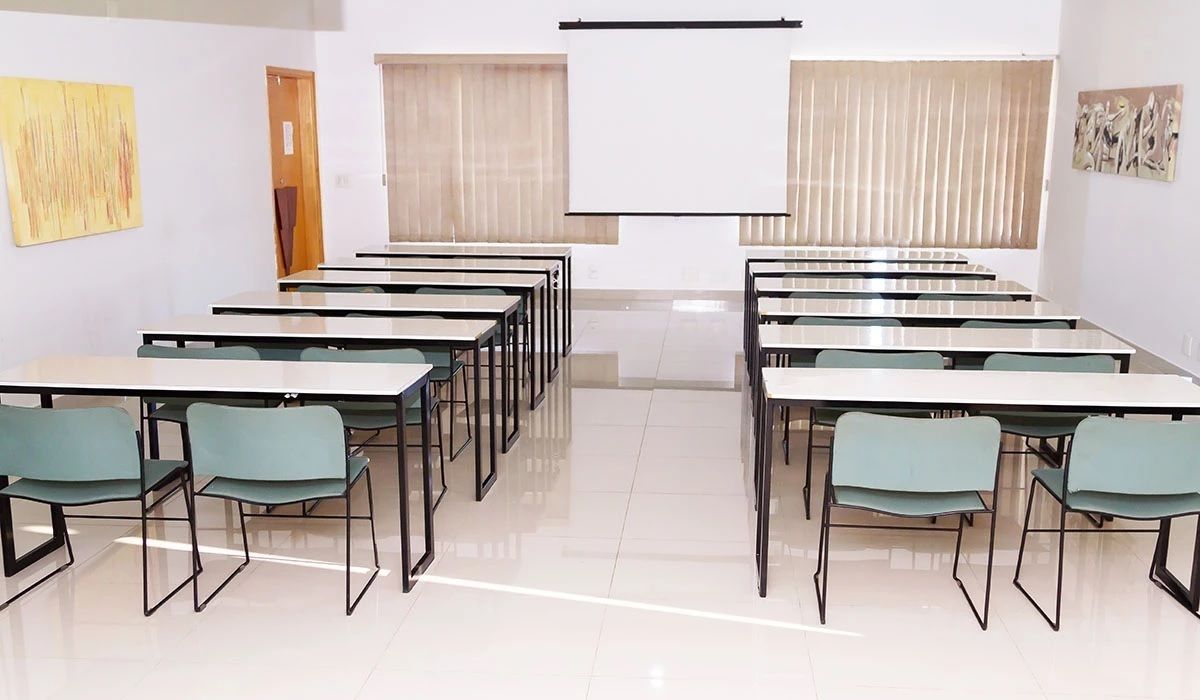 Un aula con filas de mesas y sillas, una pantalla de proyector y ventanas con persianas.