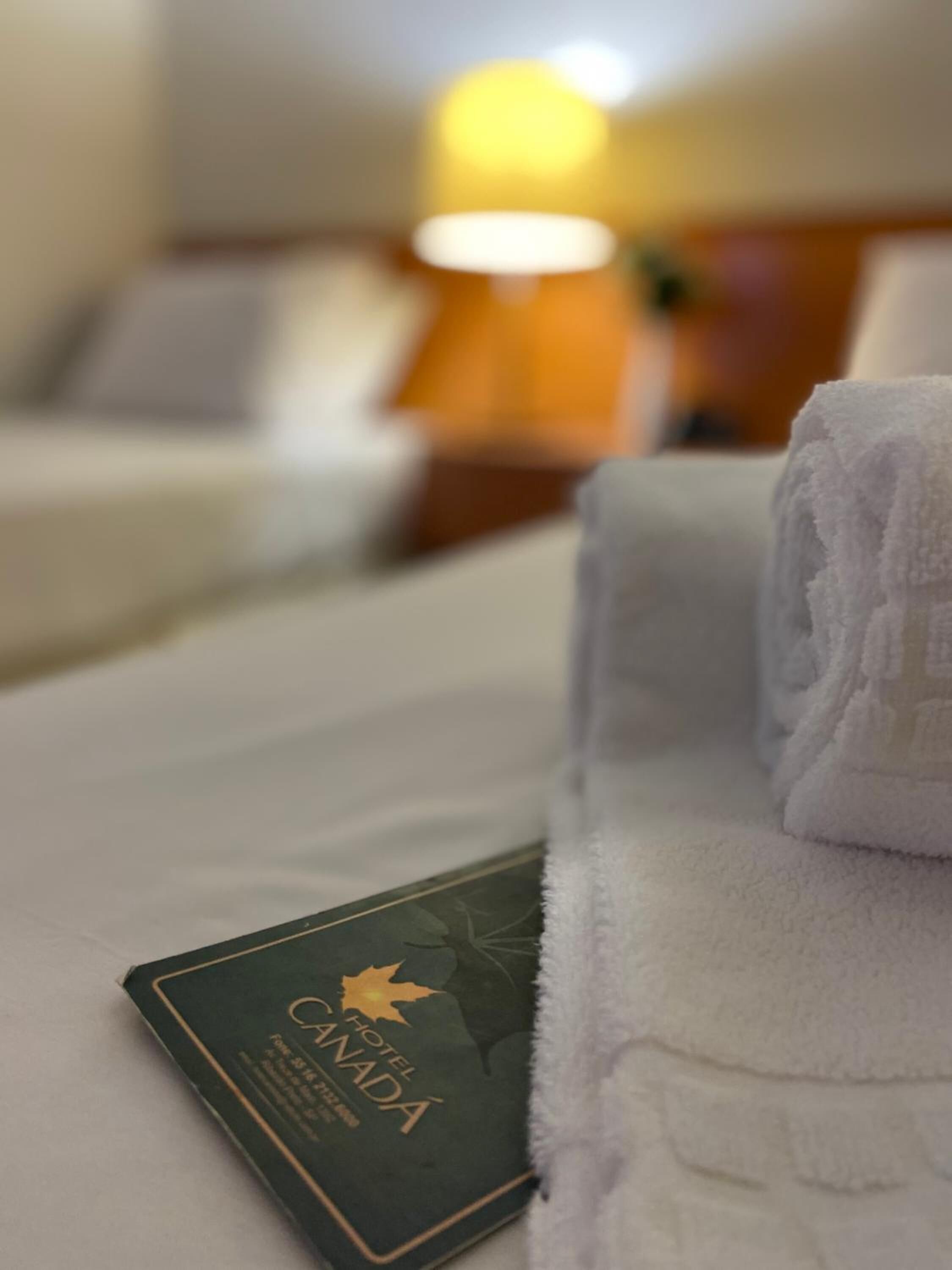A hotel room with a neatly made bed, a folded white towel, and a visible room number card.