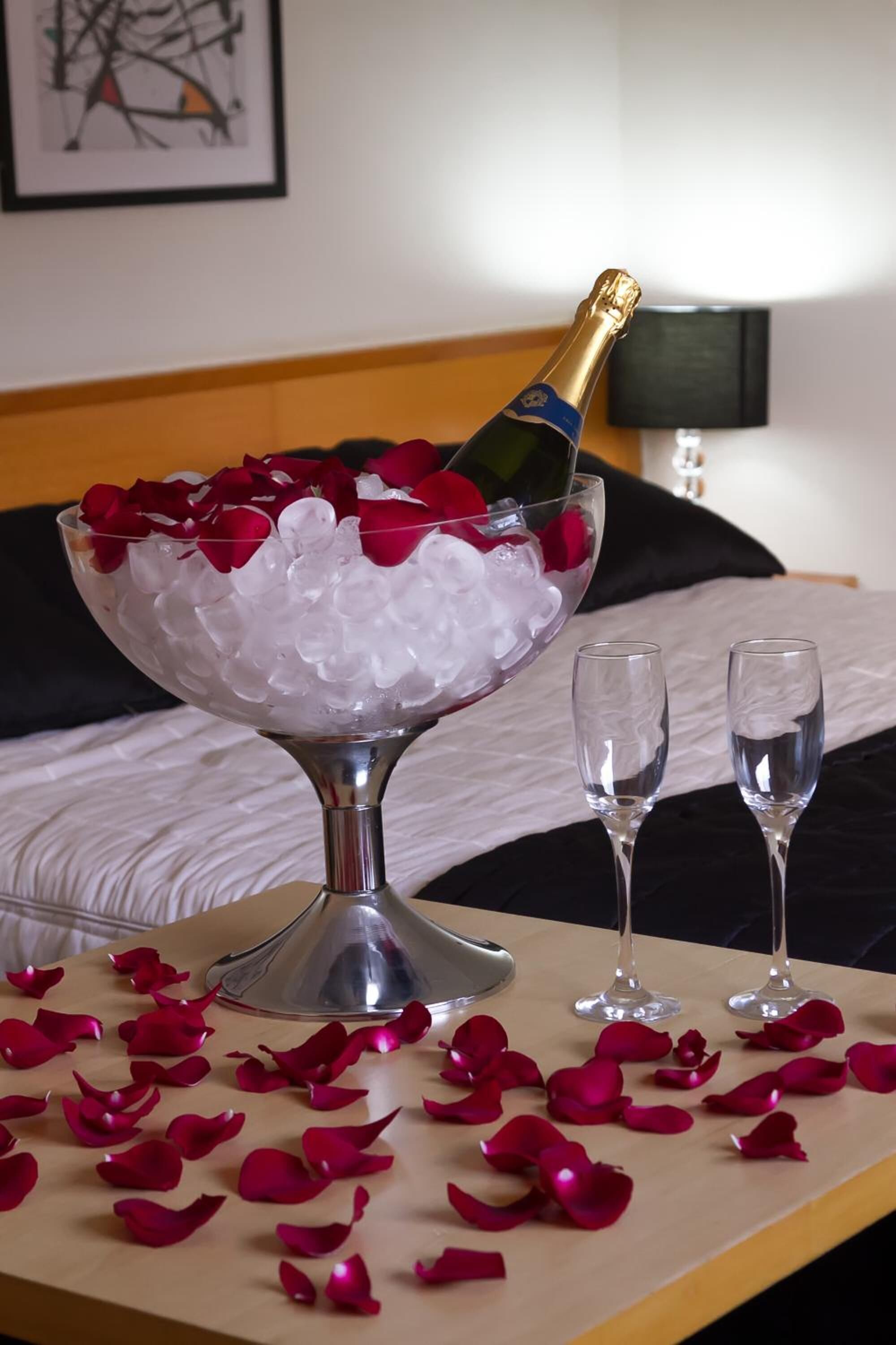 Una cama con una colcha blanca, una mesita de noche con una botella de champán, hielo y pétalos de rosa, y dos vasos vacíos sobre la mesa.