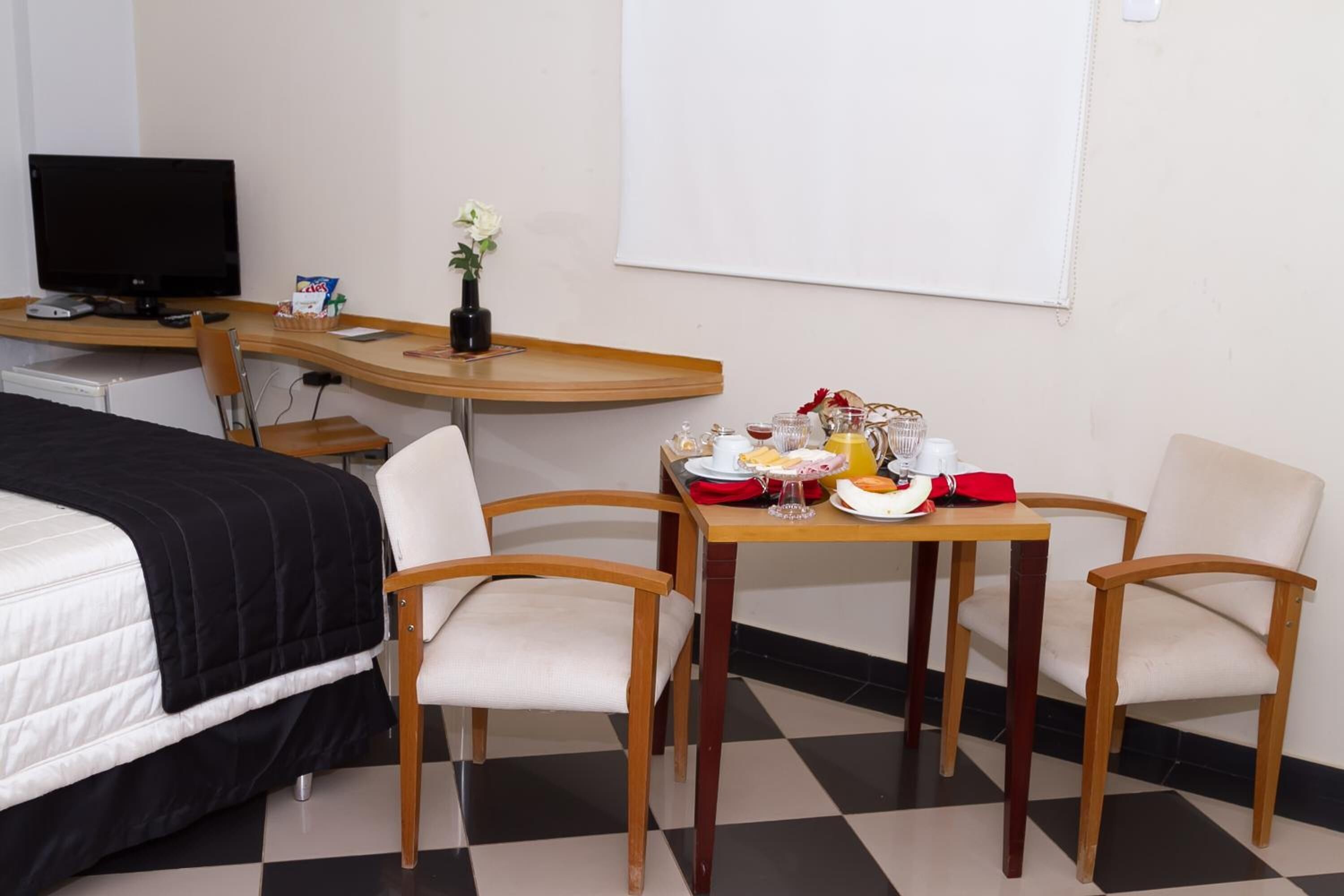 A hotel room with a bed, a desk with a computer, a dining table with chairs, and a small plant.