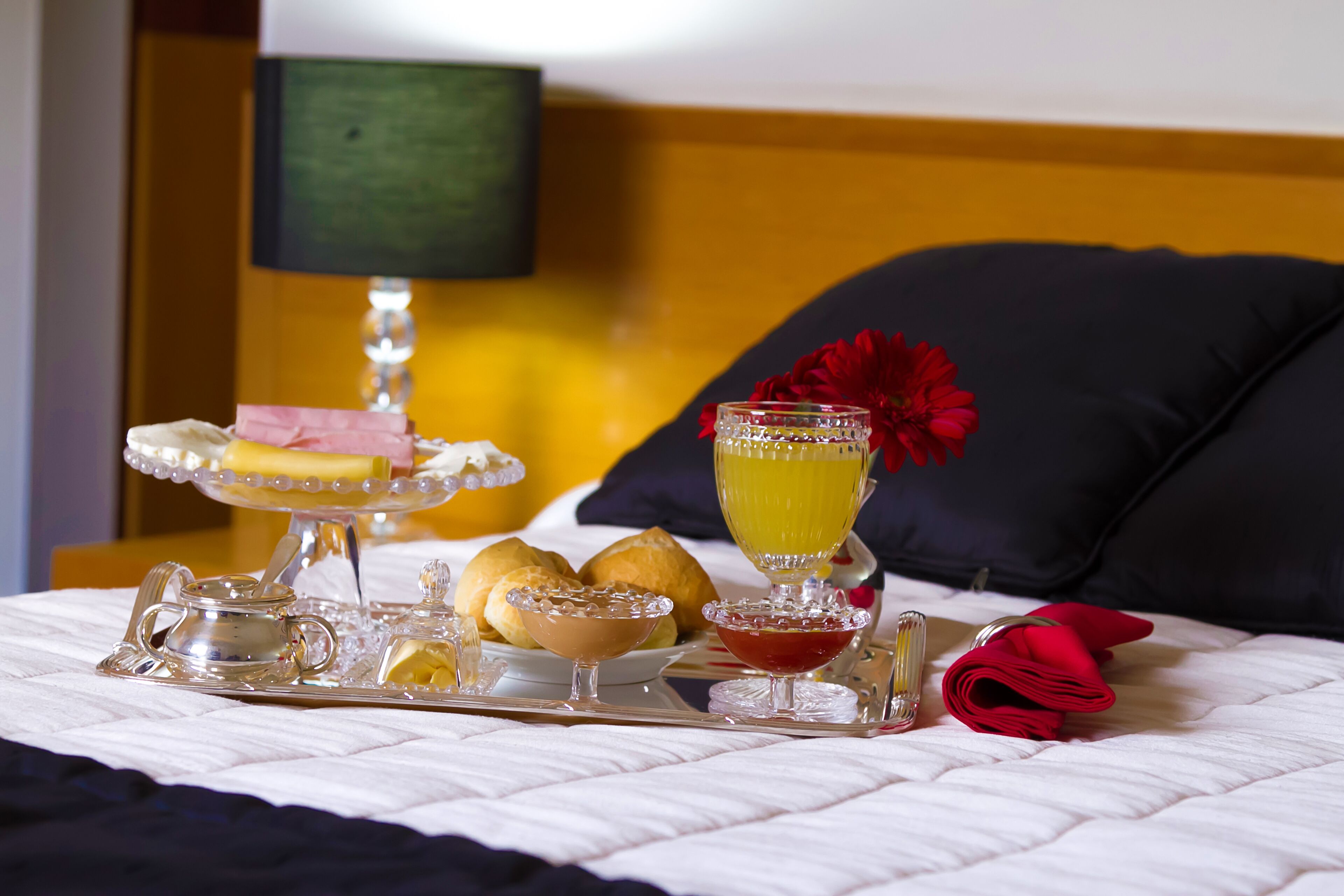 Uma bandeja de café da manhã bem organizada sobre a cama, com uma almofada e uma luminária ao fundo.